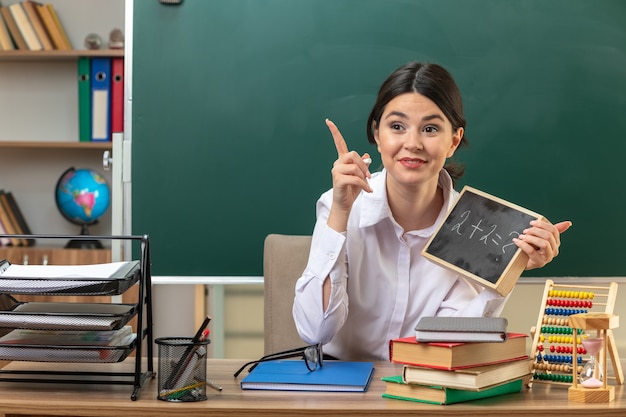 Onder de indruk van de jonge vrouwelijke leraar die aan tafel zit met schoolgereedschap met een mini-bord in de klas