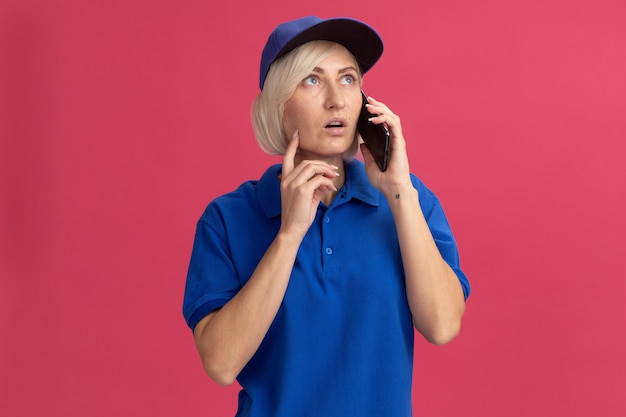 Onder de indruk van een blonde bezorger van middelbare leeftijd in blauw uniform en pet die aan de telefoon praat en haar gezicht aanraakt met de vinger