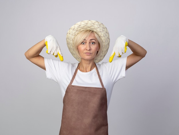 Onder de indruk van een blonde tuinmanvrouw van middelbare leeftijd in uniform met een hoed en tuinhandschoenen die naar beneden wijst, geïsoleerd op een witte muur