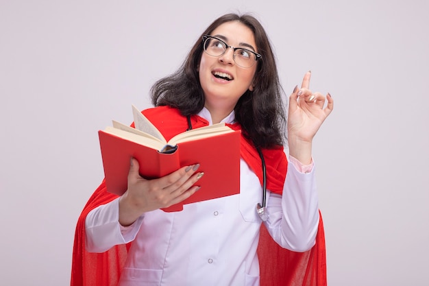 Onder de indruk van een jong kaukasisch superheldenmeisje in een rode cape met een doktersuniform en een stethoscoop met een bril die omhoog kijkt en omhoog wijst