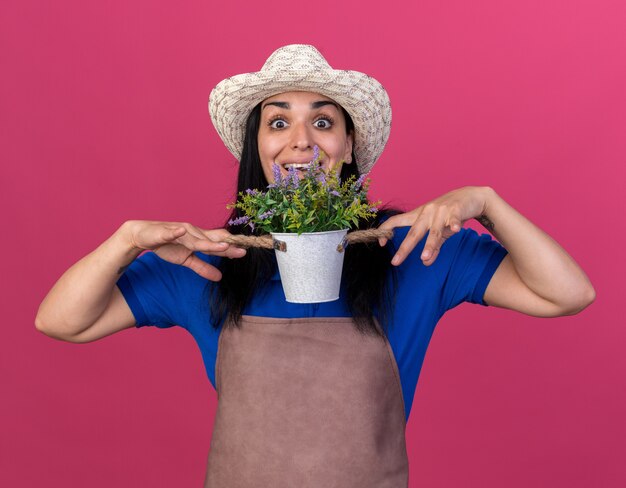 Onder de indruk van een jong kaukasisch tuinmanmeisje met uniform en hoed met bloempot geïsoleerd op roze muur