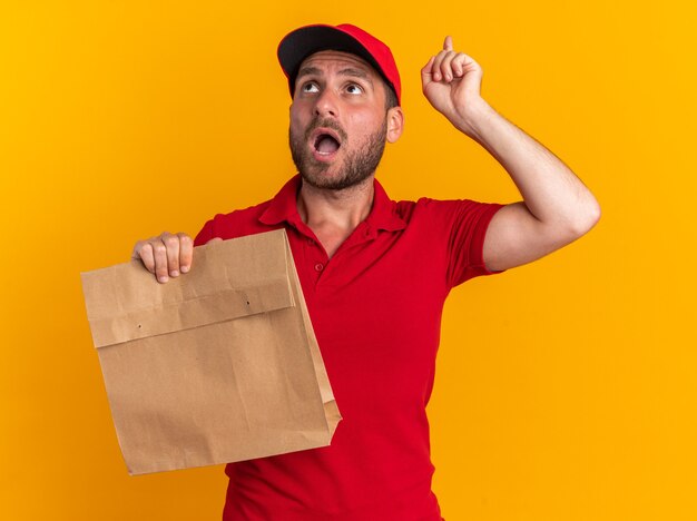 Onder de indruk van een jonge blanke bezorger in een rood uniform en een pet die een papieren pakket vasthoudt en naar boven wijst, geïsoleerd op een oranje muur