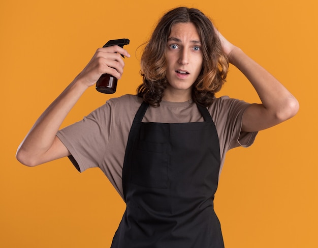 Onder de indruk van een jonge knappe kapper met een uniforme spuitfles die de hand op het hoofd houdt en naar de voorkant kijkt die op een oranje muur is geïsoleerd