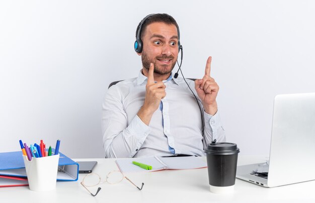 Onder de indruk van een jonge mannelijke callcentermedewerker met een headset die aan tafel zit met kantoorhulpmiddelen die naar laptoppunten kijken die omhoog geïsoleerd op een witte muur
