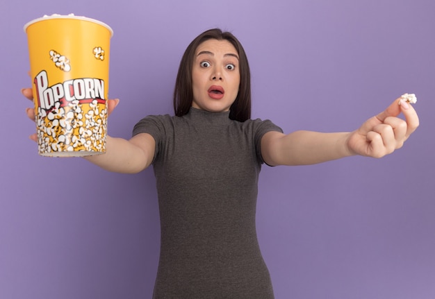 Onder de indruk van een jonge mooie vrouw die naar voren kijkt en een emmer popcorn en een stuk popcorn naar voren uitrekt, geïsoleerd op een paarse muur