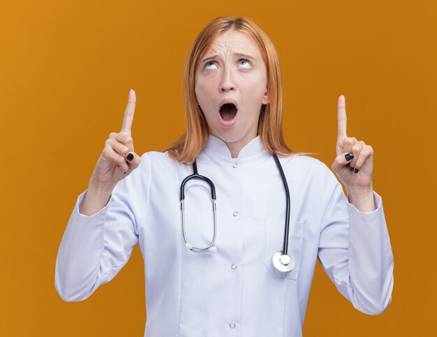 Onder de indruk van een jonge vrouwelijke gemberdokter die een medisch gewaad en een stethoscoop draagt en naar boven wijst, geïsoleerd op een oranje muur