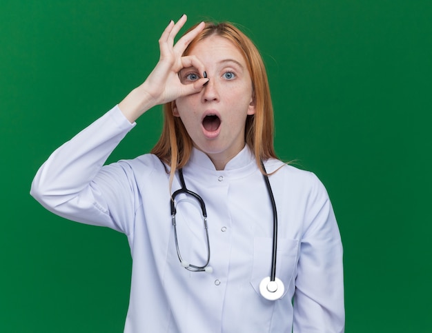 Onder de indruk van een jonge vrouwelijke gemberdokter met een medisch gewaad en een stethoscoop die naar de voorkant kijkt en een gebaar doet dat geïsoleerd is op een groene muur