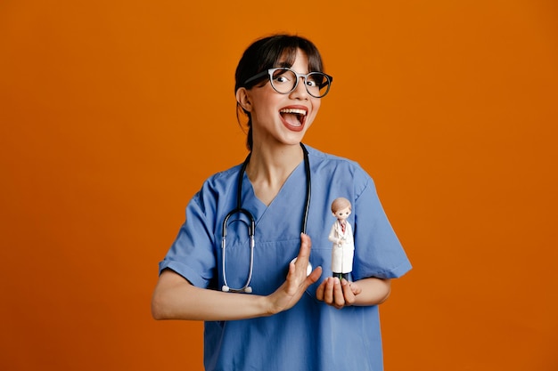 Onder de indruk van het houden van speelgoed jonge vrouwelijke arts met een uniforme stethoscoop geïsoleerd op een oranje achtergrond