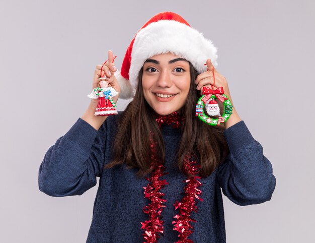 Onder de indruk van het jonge blanke meisje met kerstmuts en slinger om de nek houdt kerstboomspeelgoed geïsoleerd op een witte muur met kopieerruimte