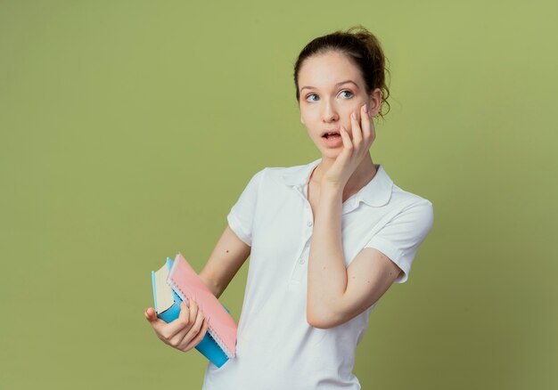 Onder de indruk van het jonge mooie vrouwelijke boek van de studentenholding en notitieblok die hand op gezicht zetten en kant bekijken die op olijfgroene achtergrond met exemplaarruimte wordt geïsoleerd