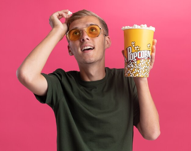 Onder de indruk van het opzoeken van jonge knappe kerel die een groen shirt en een bril draagt en een emmer popcorn houdt en een stuk popcorn op het hoofd houdt dat op roze muur wordt geïsoleerd