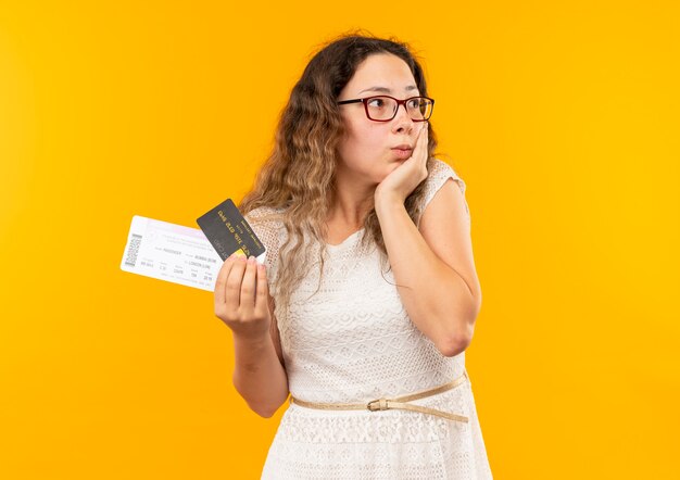 Onder de indruk van jong mooi schoolmeisje die glazen en rugtas dragen die kaartje en creditcard houden die hand op gezicht zetten die kant bekijken die op geel wordt geïsoleerd