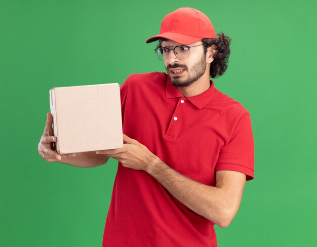 Onder de indruk van jonge bezorger in rood uniform en pet met een bril die een kartonnen doos vasthoudt en kijkt die op een groene muur is geïsoleerd