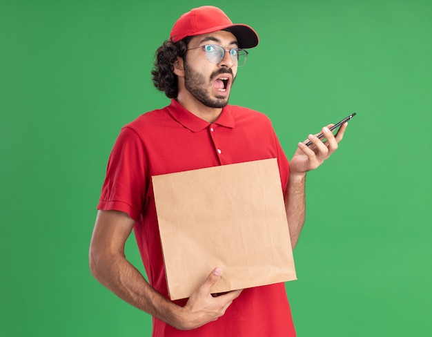 Onder de indruk van jonge blanke bezorger in rood uniform en pet met bril met papieren pakket en mobiele telefoon geïsoleerd op groene muur