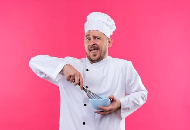 Onder de indruk van jonge knappe kok in uniform van de chef-kok die kom houdt en zwaait geïsoleerd op roze muur