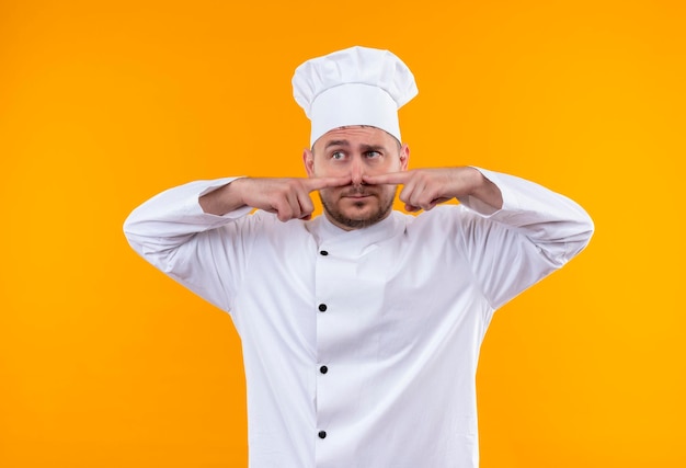 Onder de indruk van jonge knappe kok in uniform van de chef-kok die vingers op de neus zet en naar de kant kijkt die op een oranje muur is geïsoleerd