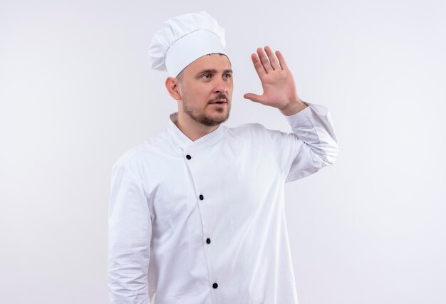 Onder de indruk van jonge knappe kok in uniform van de chef-kok kijkend naar kant met opgeheven hand geïsoleerd op een witte muur