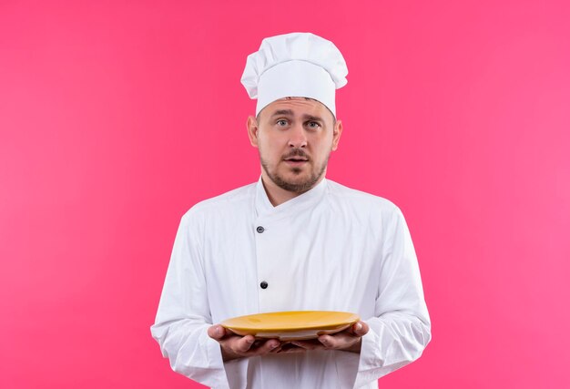 Onder de indruk van jonge knappe kok in uniform van de chef-kok met bord geïsoleerd op roze muur