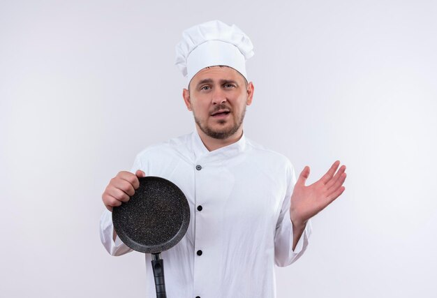 Onder de indruk van jonge knappe kok in uniform van de chef-kok met koekenpan met lege hand geïsoleerd op een witte muur