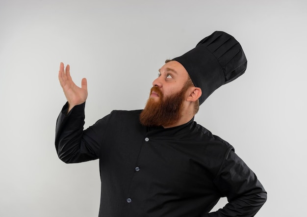 Onder de indruk van jonge knappe kok in uniform van de chef-kok met lege hand en geïsoleerd op een witte muur opzoeken