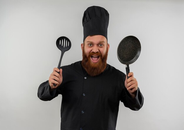 Onder de indruk van jonge knappe kok in uniform van de chef-kok met schuimspaan en koekenpan geïsoleerd op een witte muur