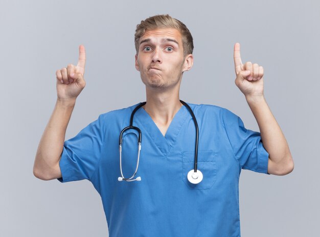 Onder de indruk van jonge mannelijke arts die doktersuniform draagt met een stethoscoop wijst naar omhoog geïsoleerd op een witte muur white