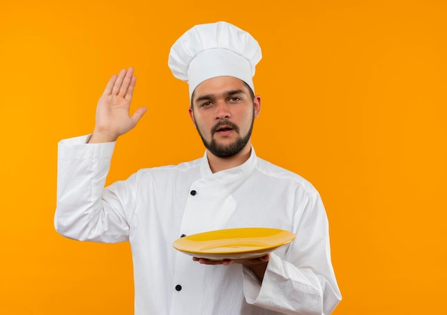 Onder de indruk van jonge mannelijke kok in uniform van de chef-kok die een leeg bord vasthoudt en de hand opsteekt geïsoleerd op een oranje muur met kopieerruimte