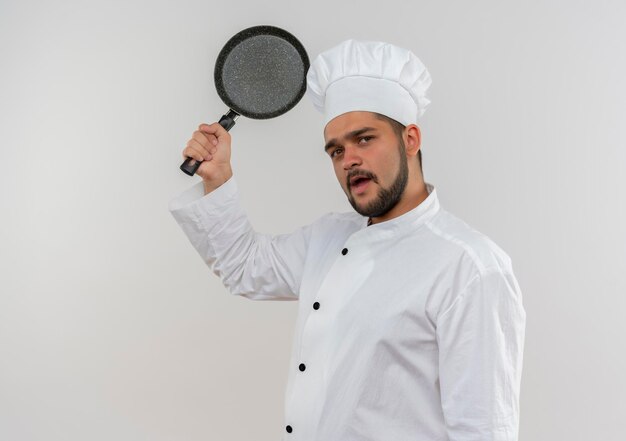 Onder de indruk van jonge mannelijke kok in uniform van de chef-kok die koekenpan op een witte muur met kopieerruimte