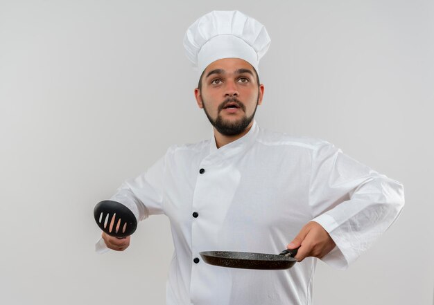 Onder de indruk van jonge mannelijke kok in uniform van de chef-kok met een lepel met sleuven en een koekenpan die op zoek is naar geïsoleerd op een witte muur