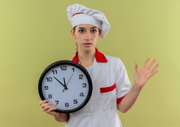 Onder de indruk van jonge mooie kok in chef-kok uniform met klok en lege hand geïsoleerd op groene muur tonen