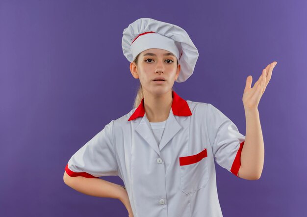 Onder de indruk van jonge mooie kok in uniform van de chef-kok die de hand opsteekt en een andere op de taille zet, geïsoleerd op de paarse muur