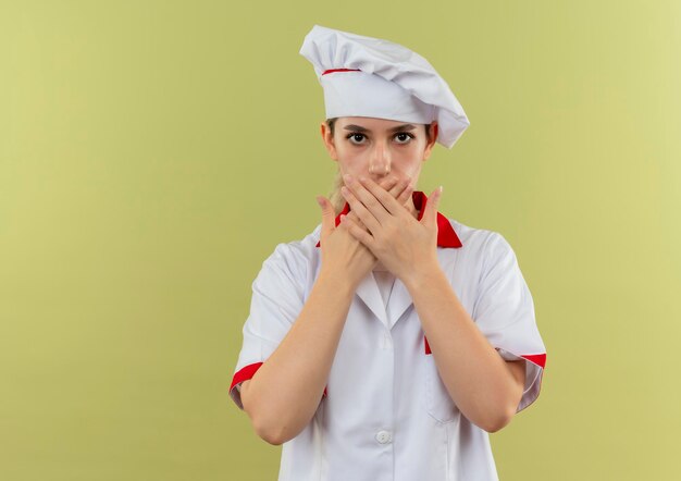 Onder de indruk van jonge mooie kok in uniform van de chef-kok die mond sluit met handen geïsoleerd op groene muur met kopieerruimte