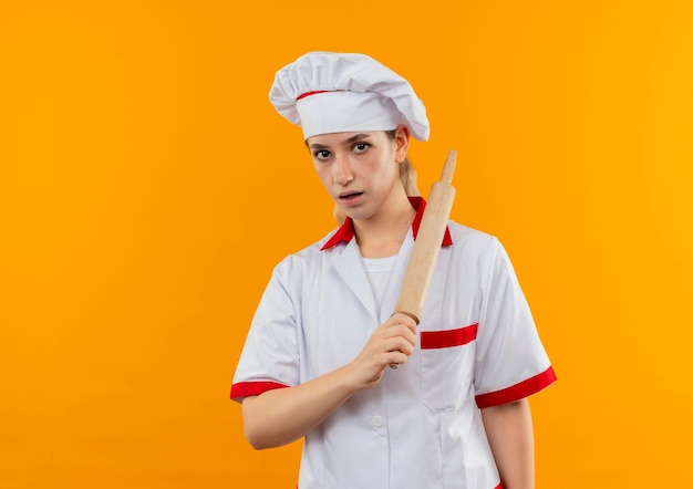 Onder de indruk van jonge mooie kok in uniform van de chef-kok met deegroller geïsoleerd op oranje muur met kopieerruimte copy