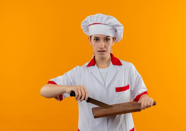 Onder de indruk van jonge mooie kok in uniform van de chef-kok met mes en snijplank geïsoleerd op oranje muur