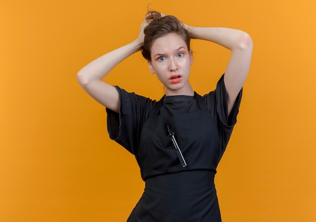 Onder de indruk van jonge Slavische vrouwelijke kapper die eenvormig holdingshoofd draagt dat op oranje achtergrond met exemplaarruimte wordt geïsoleerd