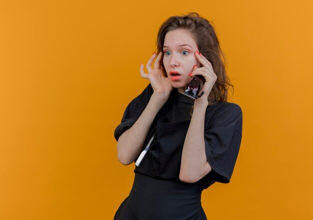 Onder de indruk van jonge Slavische vrouwelijke kapper die uniform draagt en er recht uitziet met tondeuses en gezicht aan te raken