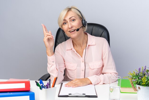 onder de indruk van jonge vrouwelijke callcentermedewerker die een headset draagt die aan tafel zit met office-tools wijst naar omhoog geïsoleerd op een witte muur