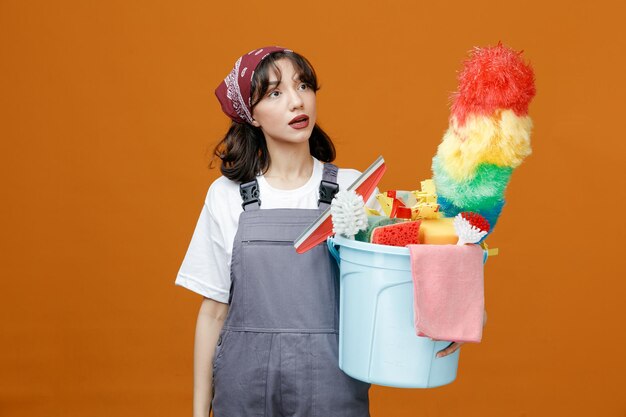 Onder de indruk van jonge vrouwelijke schoonmaker die uniform en bandana draagt met emmer met schoonmaakhulpmiddelen die naar de zijkant kijken geïsoleerd op een oranje achtergrond