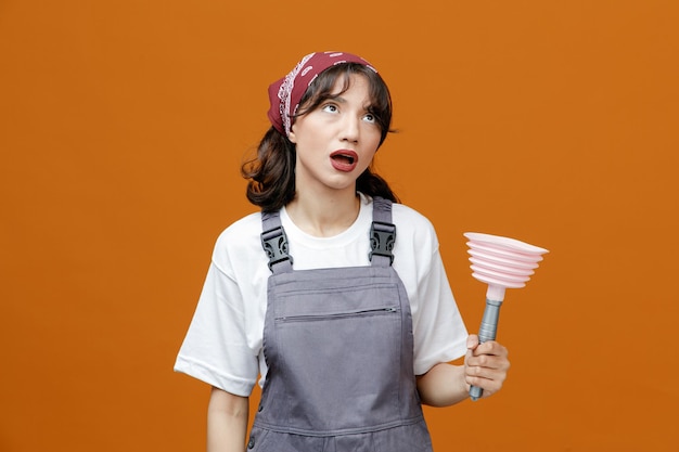 Onder de indruk van jonge vrouwelijke schoonmaker in uniform en bandana die een zuiger vasthoudt en omhoog kijkt geïsoleerd op een oranje achtergrond