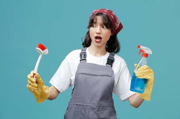 Onder de indruk van jonge vrouwelijke schoonmaker met uniforme bandana en rubberen handschoenen met borstel en reinigingsmiddel kijkend naar borstel geïsoleerd op blauwe achtergrond