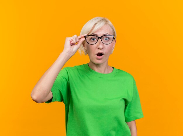 Onder de indruk van middelbare leeftijd blonde Slavische vrouw die een bril draagt die hen grijpt die naar voorzijde kijkt die op gele muur met exemplaarruimte wordt geïsoleerd