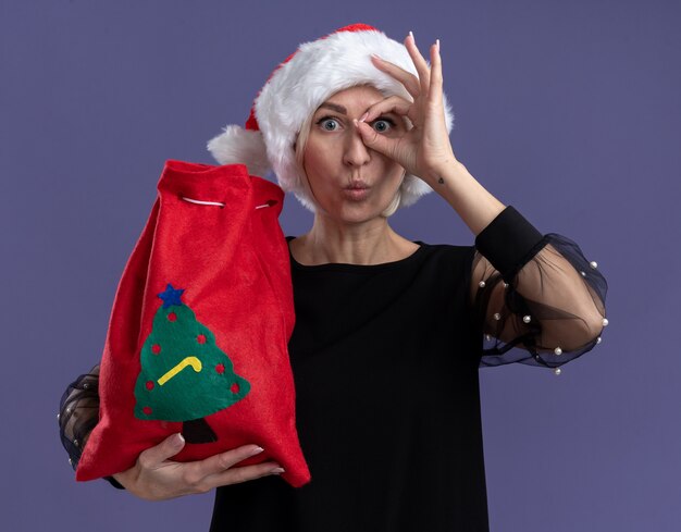 Onder de indruk van middelbare leeftijd blonde vrouw met kerstmuts bedrijf kerst zak kijken camera doen blik gebaar met samengeknepen lippen geïsoleerd op paarse achtergrond