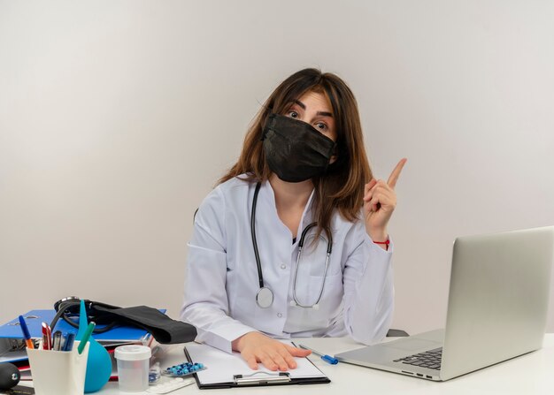 Onder de indruk van middelbare leeftijd vrouwelijke arts die medische masker draagt ?? aan bureau met medische hulpmiddelen Klembord en laptop verhogen vinger zetten hand op klembord geïsoleerd