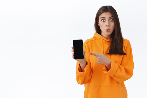 Onder de indruk van mooie jonge vrouw in oranje hoodie die lippen vouwt die tegen je praat en naar het display van de mobiele telefoon wijst om te laten zien dat de geweldige app van een vriend iets interessants online heeft gevonden om nieuws met je te delen