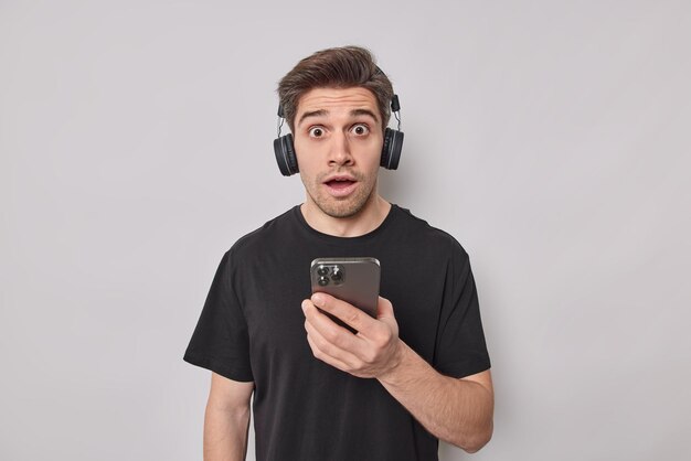 Onder de indruk verbaasde man staart afgeluisterd ogen op camera gebruikt mobiele telefoon voor online chatten luistert muziek in draadloze koptelefoon gekleed terloops geïsoleerd op witte achtergrond. Omg-concept.
