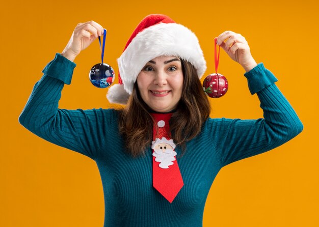 Onder de indruk volwassen blanke vrouw met kerstmuts en kerststropdas met glazen bol ornamenten geïsoleerd op oranje muur met kopie ruimte