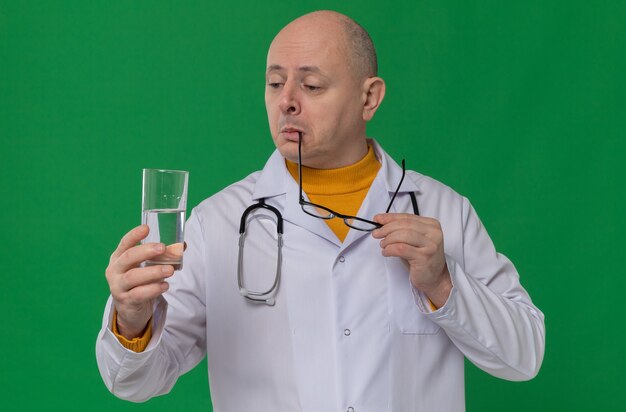 Onder de indruk volwassen Slavische man met optische bril in doktersuniform met stethoscoop die glas water vasthoudt en kijkt