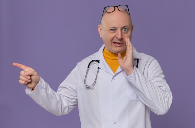 Onder de indruk volwassen slavische man met optische bril in doktersuniform met stethoscoop die hand dicht bij zijn mond houdt en naar de zijkant wijst