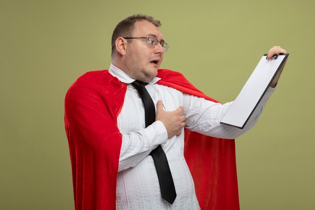 Onder de indruk volwassen Slavische superheld man in rode cape bril en stropdas vasthouden en kijken naar klembord hand op de borst zetten geïsoleerd op olijfgroene muur