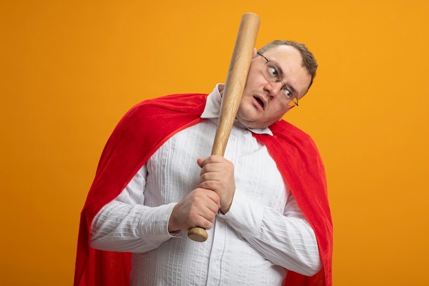 Gratis foto onder de indruk volwassen slavische superheld man in rode cape bril kijken kant zichzelf slaan in gezicht met honkbalknuppel geïsoleerd op oranje muur